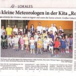 Foto von mehreren Kindern und Erwachsenen der Kita RoKoKo vor einem Gebäude. Einige halten bunte Wetter-Bastelarbeiten.