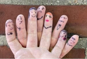 Foto einer Hand vor Backsteinwand, auf deren Fingerspitzen mit Marker verschiedene Gesichter und Accessoires gezeichnet sind.