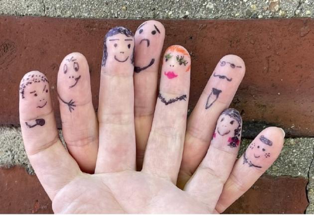 Foto einer Hand, deren Finger mit Gesichtern und Accessoires bemalt sind; Hintergrund aus Ziegelsteinen und Beton.