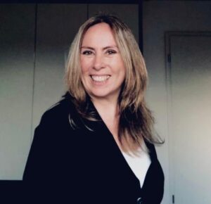Foto von Krisztina Maaß mit langen, hellbraunen Haaren, schwarzem Blazer und weißem Oberteil vor geschlossener Tür und weißen Schränken.