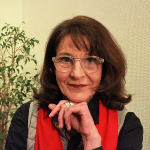 Foto von Irene Engelhardt mit schulterlangem braunem Haar, Brille und rotem Schal. Im Hintergrund eine grüne Pflanze vor heller Wand.
