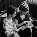 Foto von einem Mann und einer Frau auf einem Sofa. Die Frau hält ein Neugeborenes im Arm. Natürliches Licht fällt in den Raum.