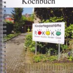 Foto des Spiral-Kochbuchs mit dem Eingang einer Kita und einem Schild "Kindertagesstätte ROKOKO Robert-Kochs-Kinder-Oase".