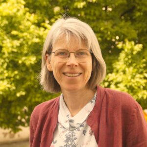 Foto von Judith Kuntz mit kurzem grauem Haar und Brille, draußen vor grünen Bäumen, im bordeauxroten Cardigan und weißer Bluse.