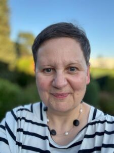 Foto von Gabriele Nemeth mit kurzem braunem Haar, blauen Augen, gestreiftem Shirt und Kette vor verschwommenem Hintergrund draußen.