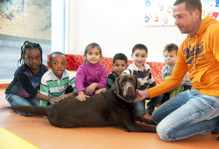 Foto von fünf Kindern und einer erwachsenen Person, die um einen großen braunen Hund sitzen und ihn in einem bunten Raum streicheln.