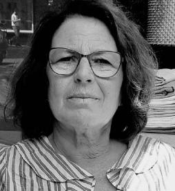 Schwarz-weiß-Foto von einer älteren Frau mit schulterlangem Haar und Brille im gestreiften Shirt, neutraler Blick in die Kamera.