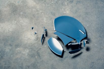 Foto von einem zerbrochenen blauen Teller, dessen große und kleine Scherben auf einer grauen, strukturierten Betonfläche liegen.